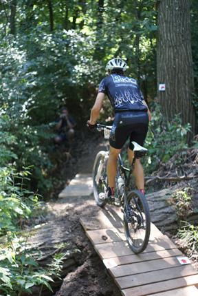 A mountain biker navigating a wooden bridge along a narrow trail in a lush, green forest. The cyclist is wearing a black jersey and shorts, and there are trees and dense foliage surrounding the path. Spanish Lake mountain bike trail.