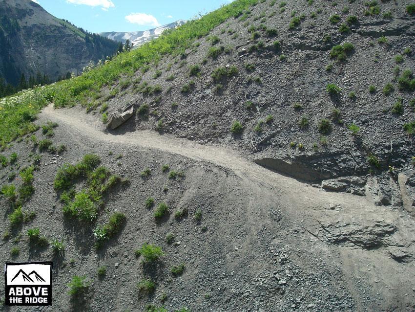 A winding dirt trail cuts through a rocky hillside, surrounded by patches of green grass and small plants. In the background, mountains are visible under a partly cloudy sky. The scene captures a natural landscape ideal for hiking and exploration. Trail 401 mountain bike trail.