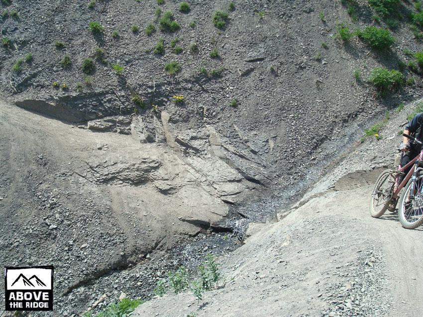 A cyclist navigating a rocky trail on a hillside, with loose gravel and sparse vegetation surrounding the path. The landscape features steep slopes and visible erosion in the terrain, highlighting the ruggedness of the area. Trail 401 mountain bike trail.