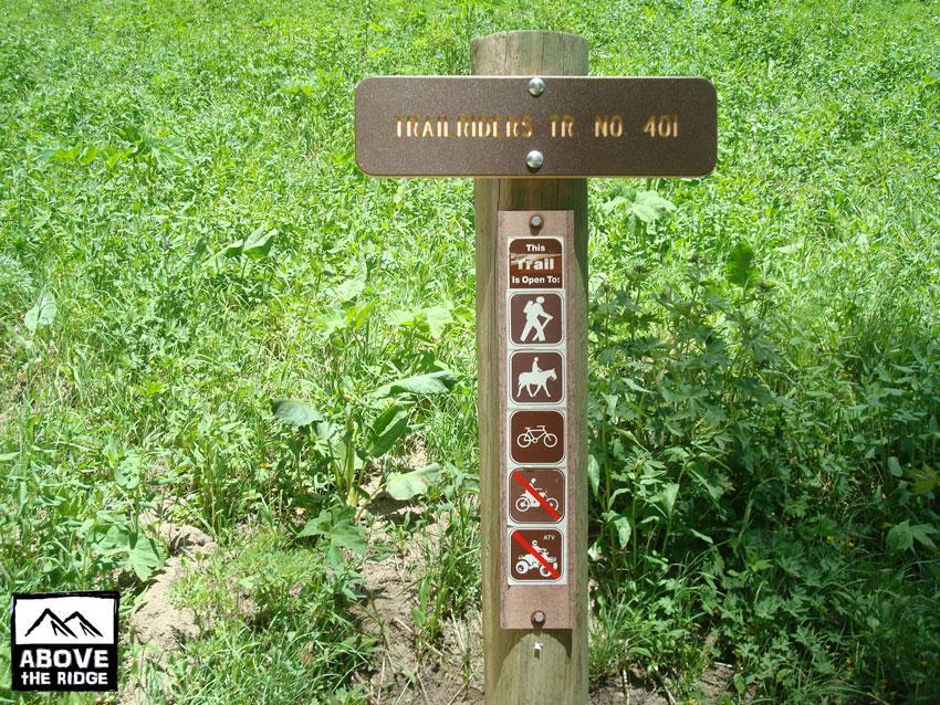 Trail sign indicating "Trail Riders Trail No. 401," featuring icons showing permitted activities such as hiking, horseback riding, and biking, with a prohibition symbol for ATVs. The sign is positioned in a grassy field. Trail 401 mountain bike trail.