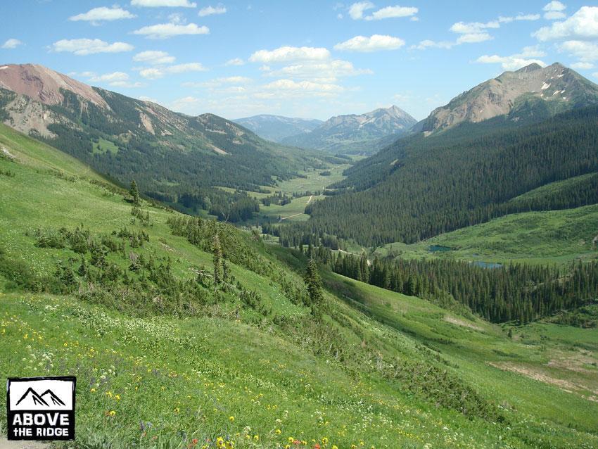 A scenic mountainous landscape featuring rolling hills covered in lush green grass and wildflowers. In the background, towering mountains stretch towards a blue sky adorned with fluffy white clouds. A valley is visible, with dense forests lining the lower areas and a winding path or river visible in the distance. The image conveys a sense of tranquility and natural beauty. Trail 401 mountain bike trail.