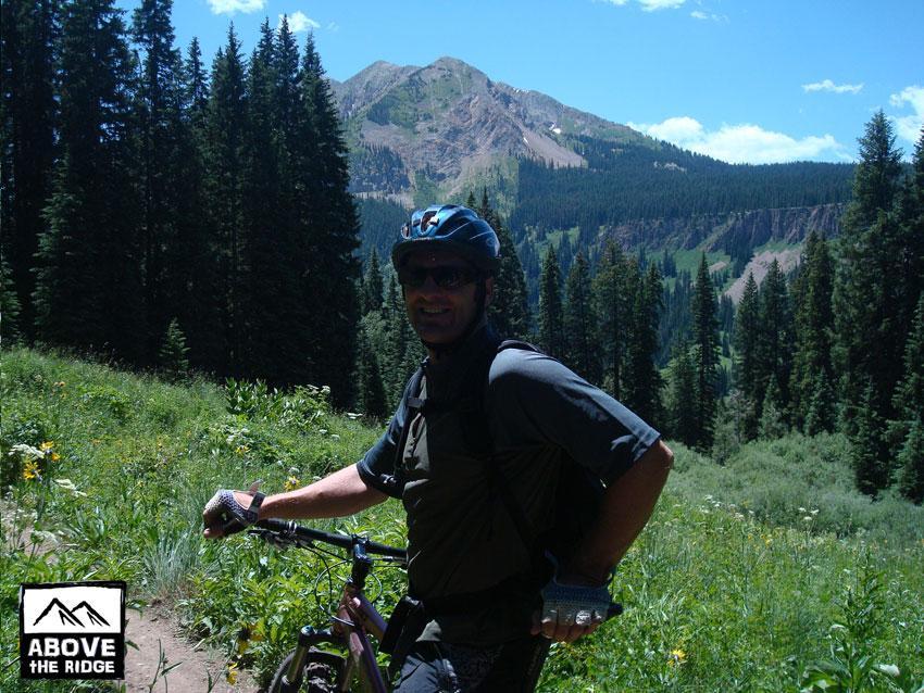 A person in a cycling helmet smiles while holding a mountain bike, standing on a trail surrounded by lush greenery and tall trees. In the background, a mountainous landscape with peaks is visible under a clear blue sky. Trail 401 mountain bike trail.