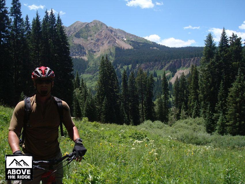 A mountain biker standing in a lush green meadow surrounded by tall pine trees, with a mountain range in the background under a clear blue sky. The cyclist is wearing a red helmet and appropriate biking gear, holding onto their bicycle. The scene captures the beauty of nature and the thrill of outdoor adventure. Trail 401 mountain bike trail.