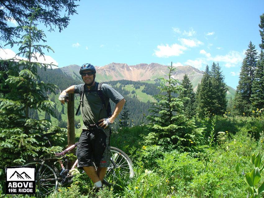 A mountain biker stands beside his bike in a lush, green landscape filled with trees and wildflowers. In the background, rolling mountains and a clear blue sky create a scenic backdrop for the outdoor adventure. The image conveys a sense of exploration and enjoyment of nature. Trail 401 mountain bike trail.