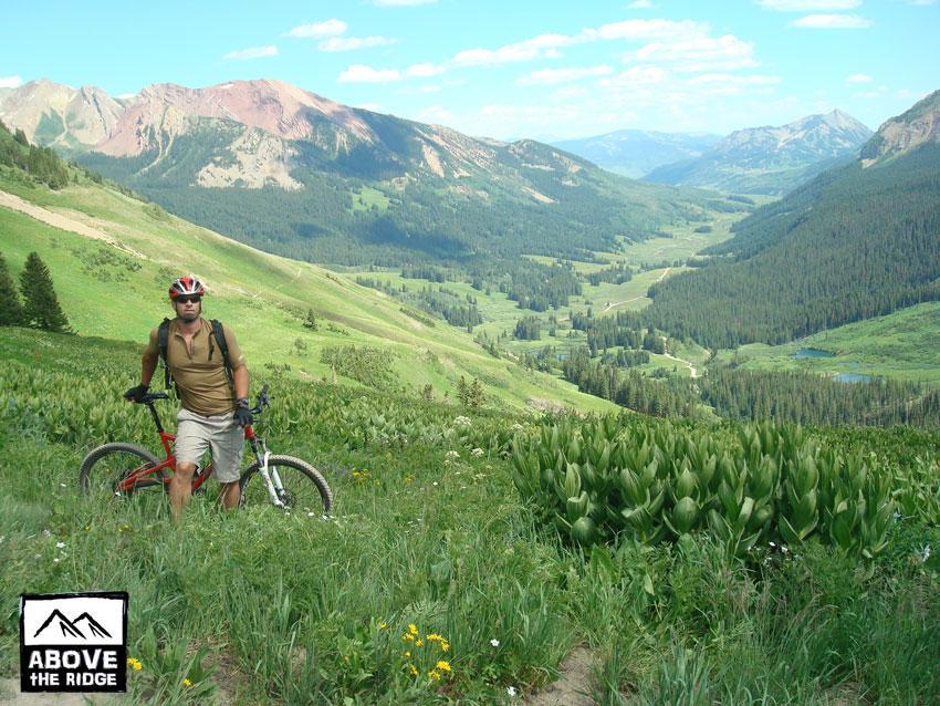 A mountain biker standing beside a bicycle in a grassy mountain landscape, with rolling hills and a lush green valley in the background under a clear blue sky. Trail 401 mountain bike trail.
