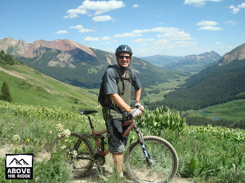 A mountain biker standing next to a bike on a lush green hillside, surrounded by mountains and valleys under a clear blue sky with scattered clouds. The biker is wearing a helmet and a sporty outfit, smiling while enjoying the scenic view. Trail 401 mountain bike trail.