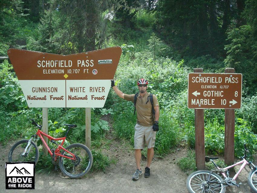 A cyclist stands next to a sign indicating Schofield Pass at an elevation of 10,707 feet, surrounded by dense greenery. A mountain bike is leaned against the sign on the left, while the right sign points to Gothic (8 miles) and Marble (10 miles). The setting is within the Gunnison and White River National Forests. Trail 401 mountain bike trail.