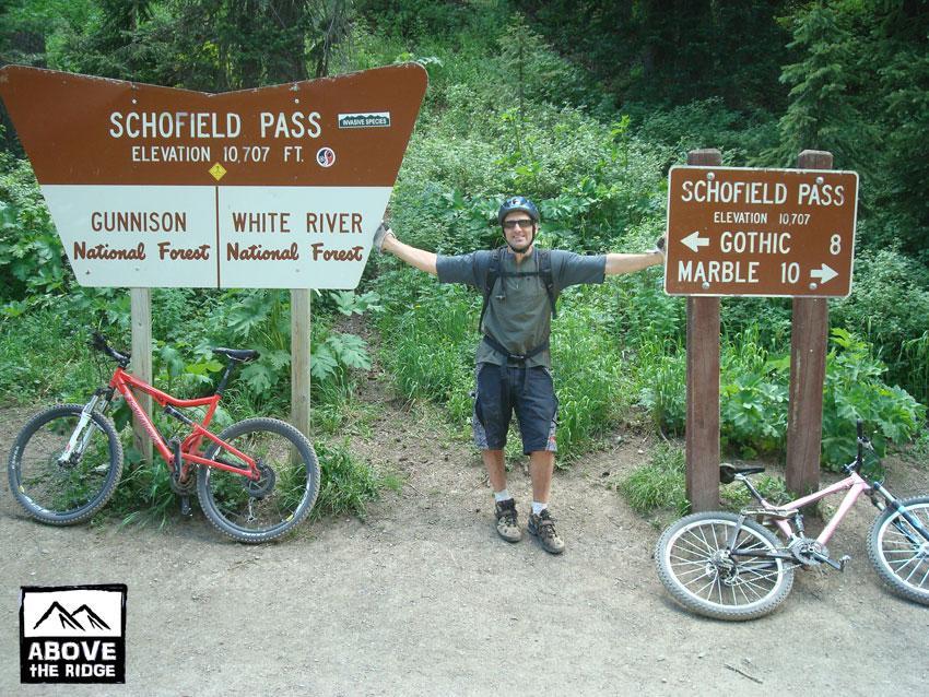 A mountain biker stands with arms outstretched at Schofield Pass, marked at an elevation of 10,707 feet. Two bicycles are parked nearby: one red and one pink. Signs indicate directions to Gothic and Marble, and the lush greenery of Gunnison and White River National Forests surrounds the area. Trail 401 mountain bike trail.