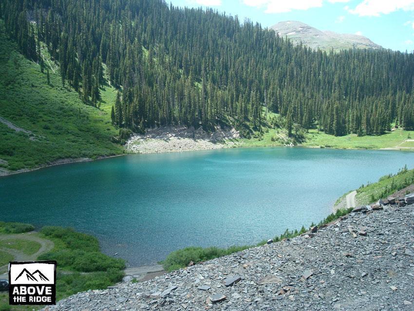 A scenic view of a turquoise lake surrounded by lush green hills and dense evergreen trees under a clear blue sky. The shoreline features rocky areas and grassy patches, creating a serene natural landscape. Trail 401 mountain bike trail.