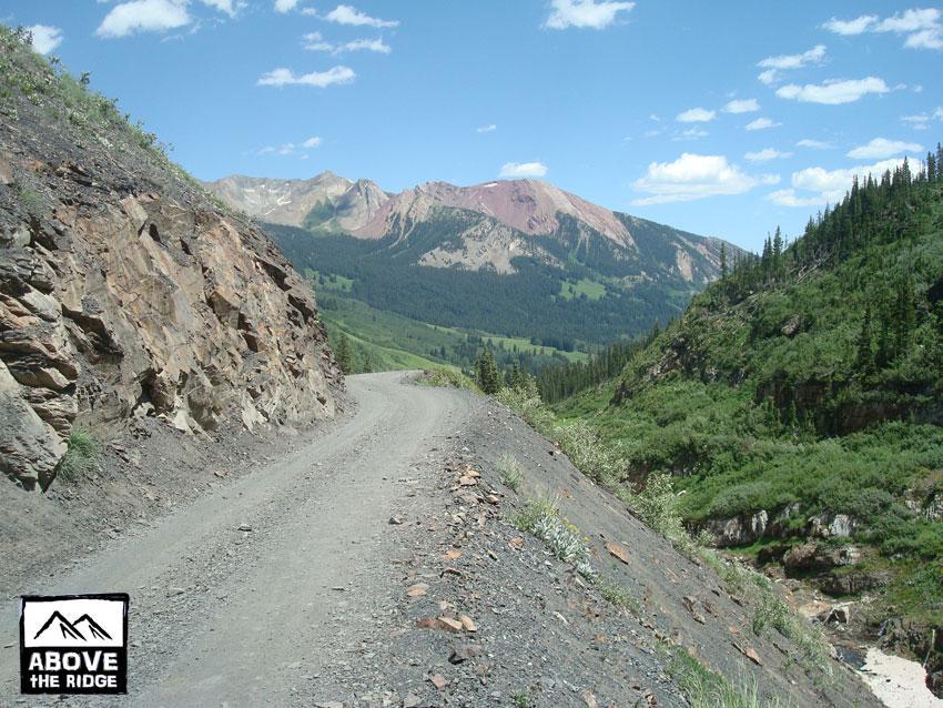A scenic dirt road winding through a mountainous landscape, with rocky cliffs on one side and lush greenery on the other. In the background, majestic mountains rise under a blue sky dotted with fluffy white clouds. A small stream can be seen meandering through the valley. Trail 401 mountain bike trail.