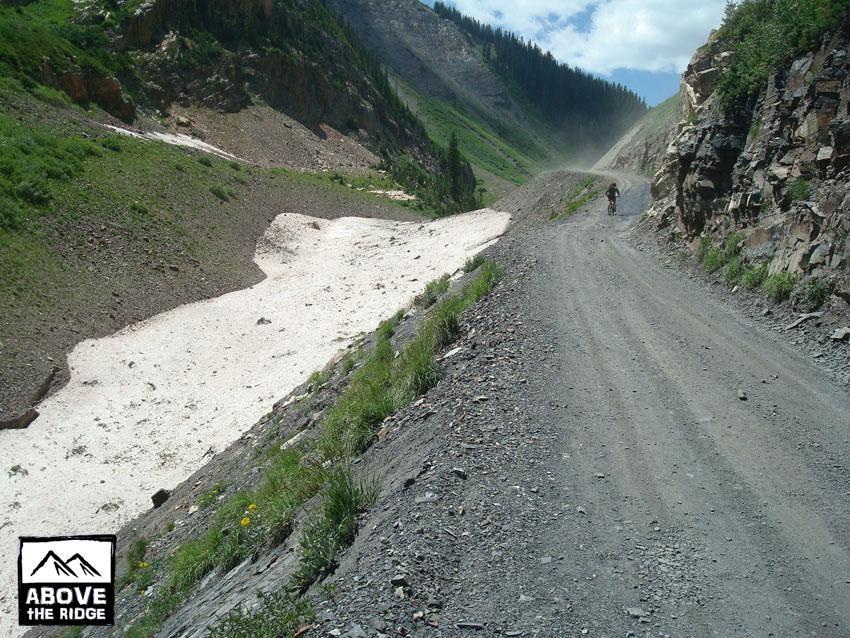 A gravel road winding through a mountainous landscape, with steep rocky cliffs on both sides and patches of green vegetation. In the foreground, a cyclist rides along the road, with a dust cloud trailing behind. In the background, there are snowy patches on the ground alongside the road, indicative of higher altitudes and cooler temperatures. The sky is partly cloudy, suggesting a sunny day. Trail 401 mountain bike trail.