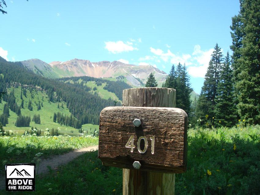 A rustic wooden signpost with the number "401" stands in a lush green landscape. The backdrop features rolling hills and mountains under a clear blue sky with scattered clouds. Pine trees adorn the scene, indicating a natural, mountainous area, possibly a hiking trail or scenic viewpoint. Trail 401 mountain bike trail.