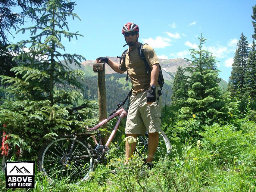 A cyclist in outdoor gear stands next to a mountain bike and a trail sign in a lush, green forest. The backdrop features mountains under a blue sky with scattered clouds. The cyclist is wearing a helmet and gloves, and the scene captures the essence of adventure and outdoor activity. Trail 401 mountain bike trail.