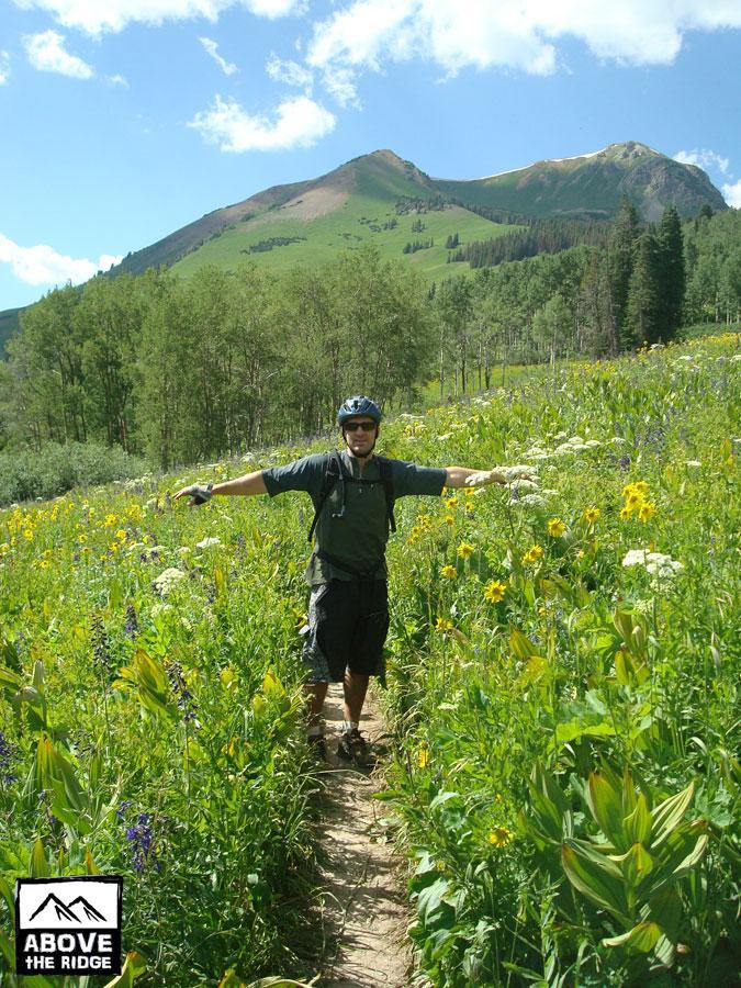 A person standing on a narrow dirt path surrounded by vibrant wildflowers in a lush green landscape. The background features a mountain with trees and blue skies dotted with clouds. The individual is wearing a helmet and cycling gear, with arms outstretched, conveying a sense of joy and adventure. Trail 401 mountain bike trail.