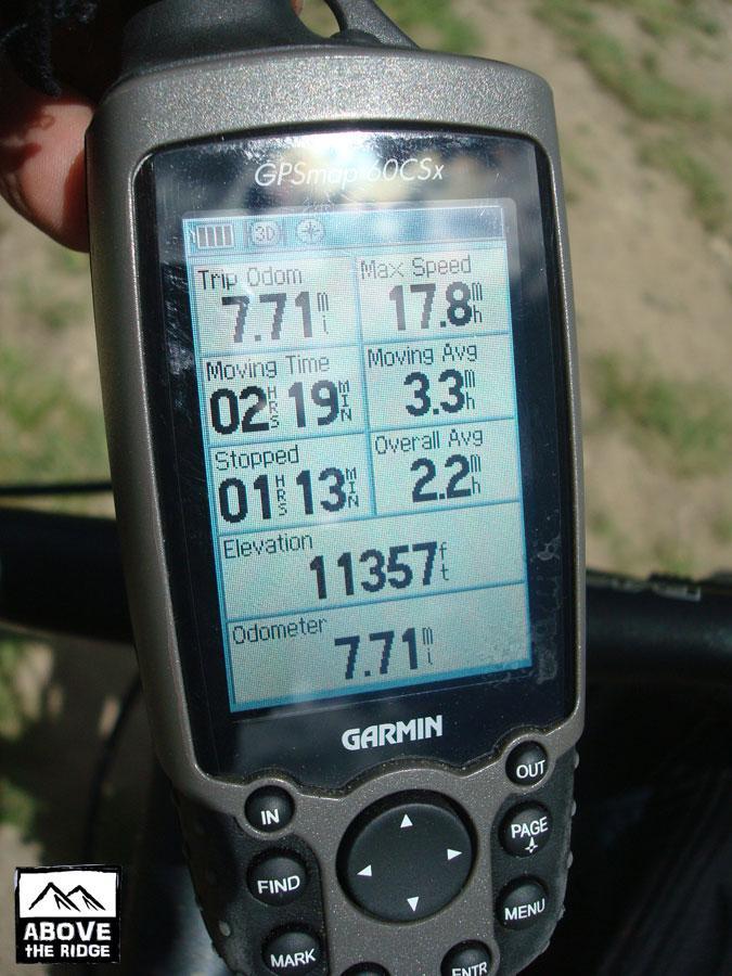 A close-up view of a Garmin GPS device displaying various metrics: trip odometer reading 7.71 miles, maximum speed of 17.8 mph, moving time of 2 hours and 19 minutes, moving average of 3.3 mph, and an elevation of 11,357 feet. The screen also shows a total stopped time of 1 minute and 13 seconds and an overall average speed of 2.2 mph. The device is being held in one hand, with a blurred outdoor background. Trail 401 mountain bike trail.