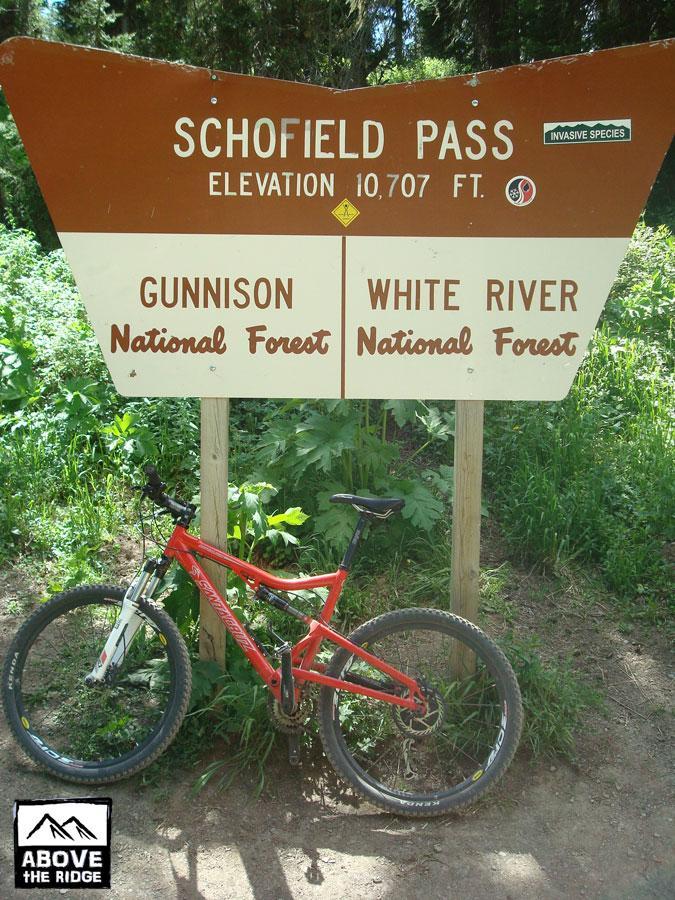 A mountain bike rests beside a sign for Schofield Pass, indicating an elevation of 10,707 feet. The sign features information about Gunnison National Forest and White River National Forest, with greenery and foliage in the background. Trail 401 mountain bike trail.