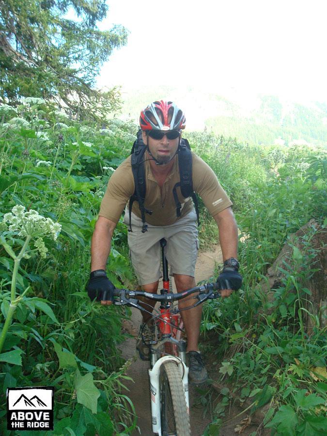 A man riding a mountain bike on a narrow dirt trail surrounded by lush greenery, wearing a helmet, gloves, and a backpack. The scene depicts an outdoor adventure in a natural setting. Trail 401 mountain bike trail.