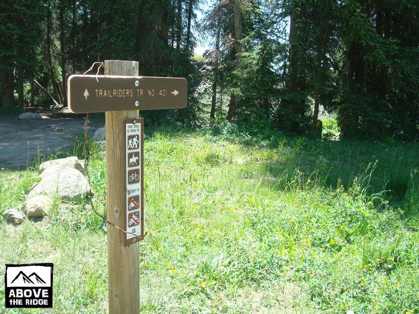 A wooden trail sign indicating "Trailriders Trail No. 401" with symbols for hiking, horseback riding, and mountain biking in a green, grassy area surrounded by trees. Trail 401 mountain bike trail.
