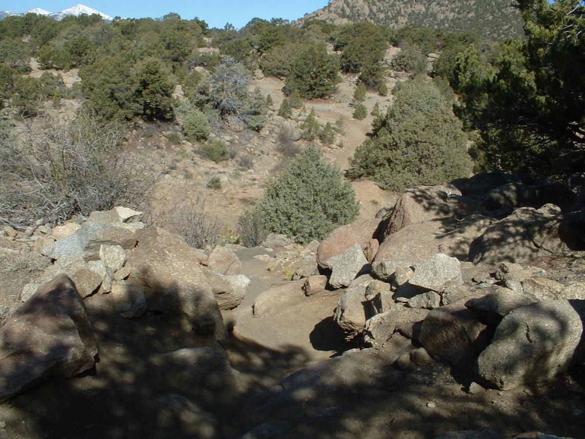 A rocky landscape featuring scattered boulders and sparse vegetation, with a backdrop of rolling hills and distant snow-capped mountains under a clear blue sky. Midland Hills Trails mountain bike trail.