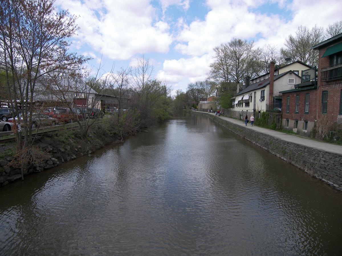 "View of a tranquil canal lined with trees and buildings, with people walking along the pathway. The water reflects the cloudy sky, and nearby parked cars can be seen." Bridge To Bridge - D&r Canal mountain bike trail.