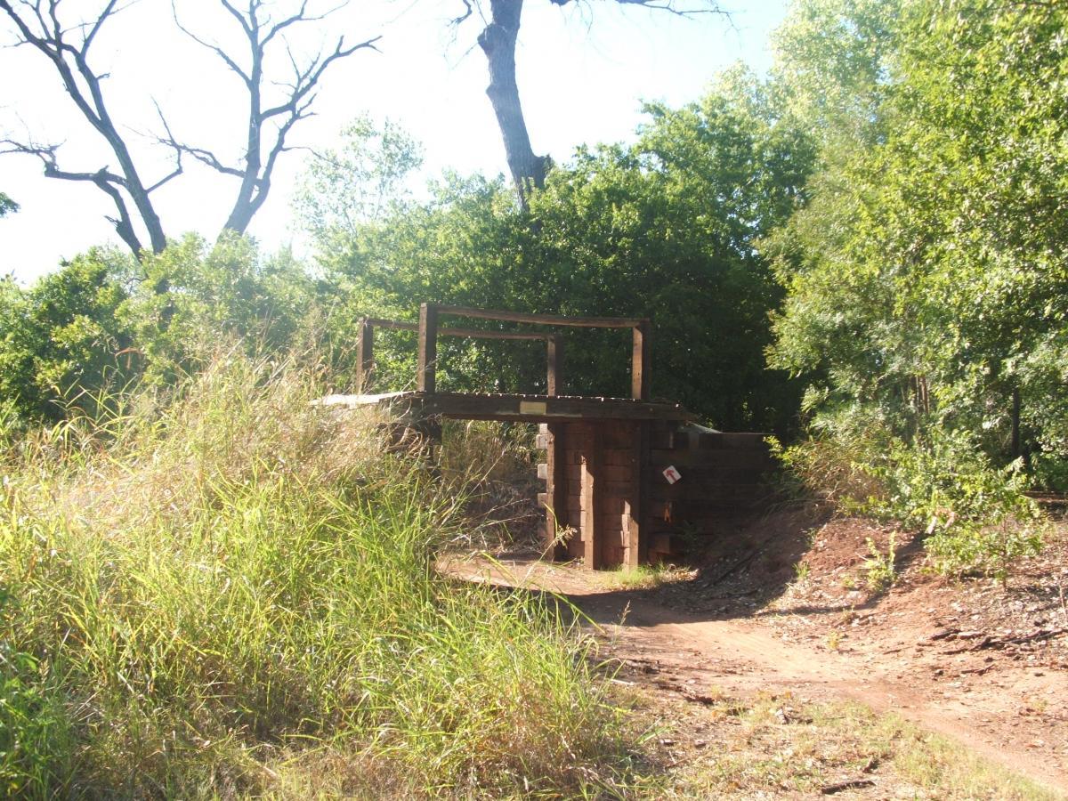 A wooden bridge situated on a dirt path, surrounded by tall grasses and trees under a bright blue sky. The bridge is elevated slightly above ground level, leading into a natural landscape. Wee-chi-ta mountain bike trail.