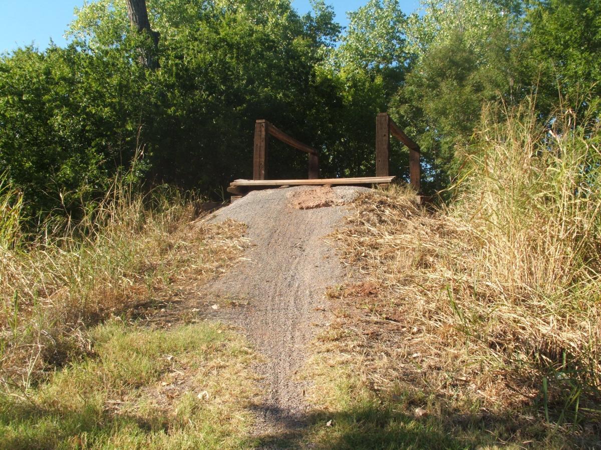 A dirt path leading up to a wooden bridge, surrounded by overgrown grass and trees. The scene is sunny and features a clear blue sky. Wee-chi-ta mountain bike trail.