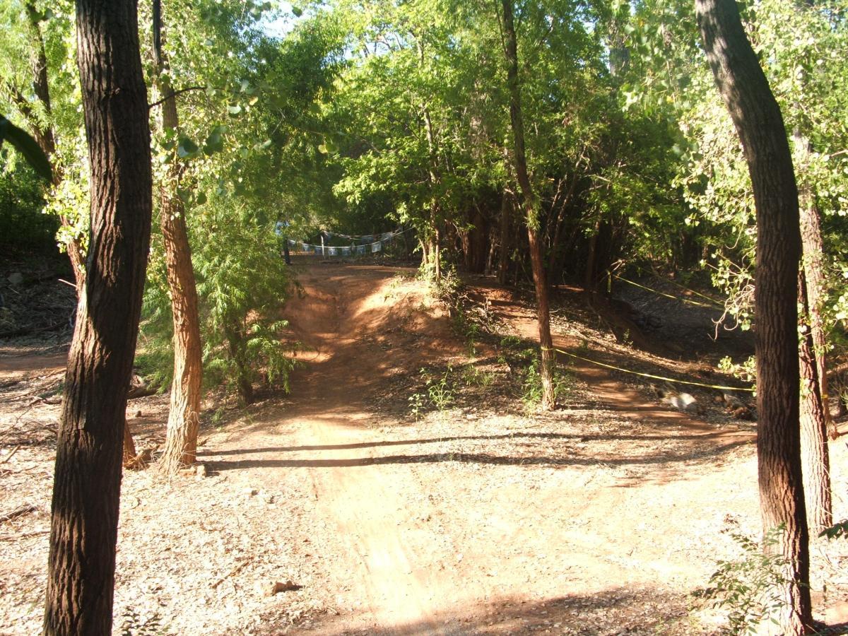 A dirt path winding through a wooded area, surrounded by tall trees and greenery. Sunlight filters through the leaves, creating dappled shadows on the ground. A small embankment is visible in the background, leading to a fenced area. Wee-chi-ta mountain bike trail.