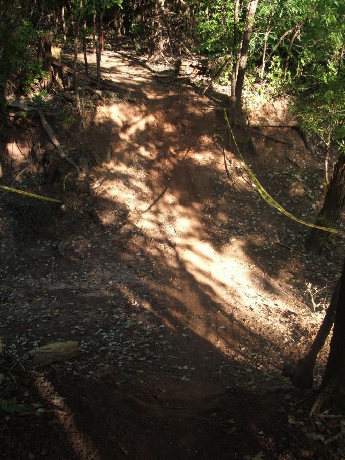 A dirt path with a steep incline, surrounded by trees and underbrush. Yellow caution tape is visible along the edge of the trail. Sunlight filters through the foliage, casting shadows on the ground. Wee-chi-ta mountain bike trail.