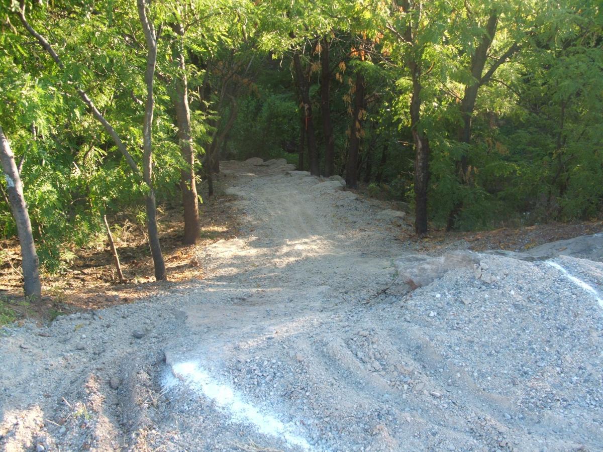 A gravel path leading into a wooded area, surrounded by trees with green foliage. The path appears to be freshly cleared and has some uneven patches, with a mound of gravel visible in the foreground. Sunlight filters through the trees, creating a serene, natural setting. Wee-chi-ta mountain bike trail.