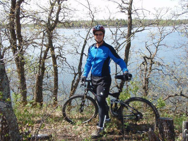 A person wearing a blue cycling jersey and helmet is standing beside a mountain bike on a hillside near a body of water, surrounded by bare trees and greenery. The scene is set on a sunny day with clear skies. Lake Metigoshe State Park mountain bike trail.