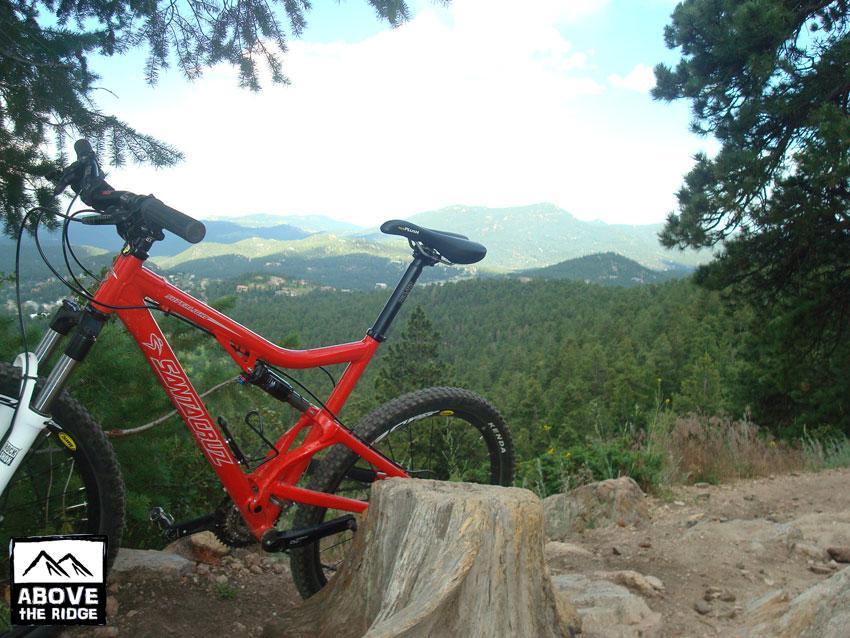 A red mountain bike leaning against a wooden stump, set against a scenic backdrop of rolling hills and pine trees. The sky is partly cloudy, highlighting the vibrant green landscape in the distance. Elk Meadow mountain bike trail.