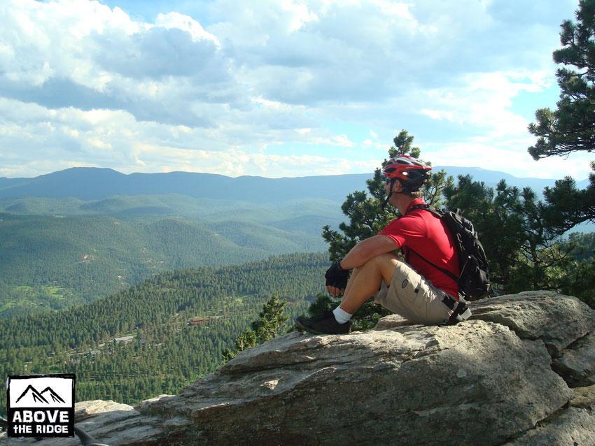 A cyclist sitting on a large rock, gazing over a lush mountain landscape under a partly cloudy sky. The person is wearing a red shirt, shorts, and a helmet, with a backpack beside them. The scene captures a serene moment in nature, showcasing the expansive view of rolling hills and greenery. Elk Meadow mountain bike trail.