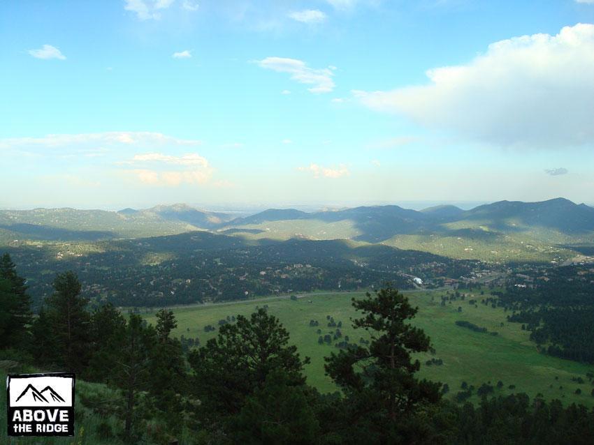 A panoramic view of rolling hills and valleys under a bright blue sky, showcasing lush green landscapes and distant mountains, with patches of clouds scattered throughout the sky. Trees are visible in the foreground, providing a natural frame to the scenery. The image is marked with the logo "Above the Ridge" in the bottom left corner. Elk Meadow mountain bike trail.