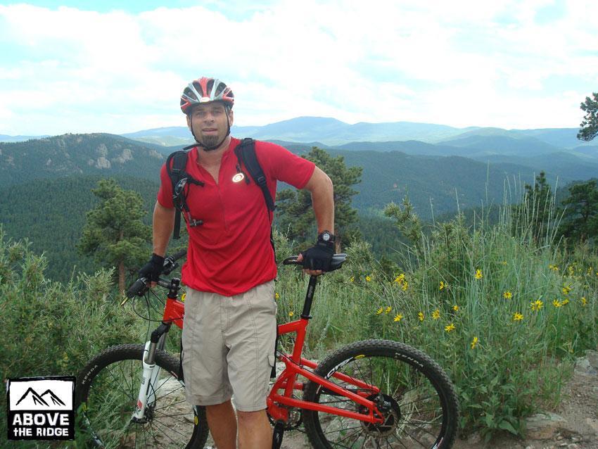 A mountain biker wearing a red shirt and helmet is standing next to a red bike on a scenic trail. The background features lush green mountains and a clear sky, with hints of wildflowers nearby. Elk Meadow mountain bike trail.
