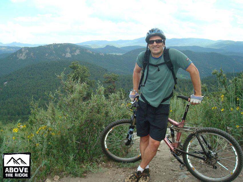 A smiling cyclist standing next to a mountain bike on a trail, with a scenic view of rolling hills and trees in the background. The cyclist is wearing a helmet, sunglasses, and casual cycling attire. Wildflowers are visible nearby, and the sky is partly cloudy. Elk Meadow mountain bike trail.