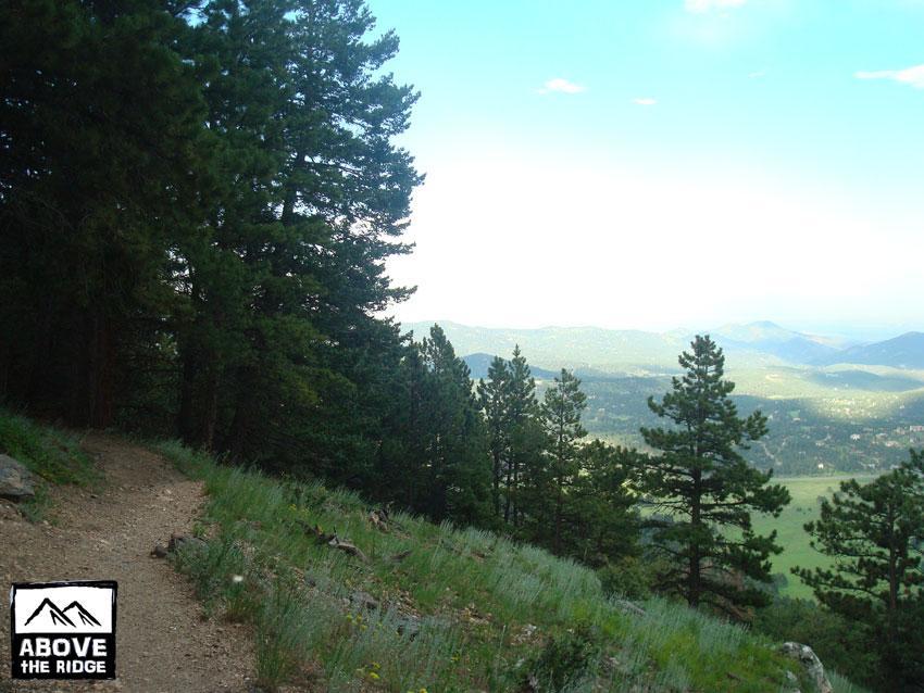 A winding dirt path surrounded by tall pine trees leads to a scenic view of valleys and distant mountains, under a partly cloudy sky. In the bottom left corner, there is a logo reading "Above the Ridge." Elk Meadow mountain bike trail.