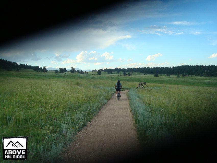 A person riding a bicycle along a dirt path through a lush green field, bordered by trees and mountains in the background, under a partly cloudy sky. Elk Meadow mountain bike trail.
