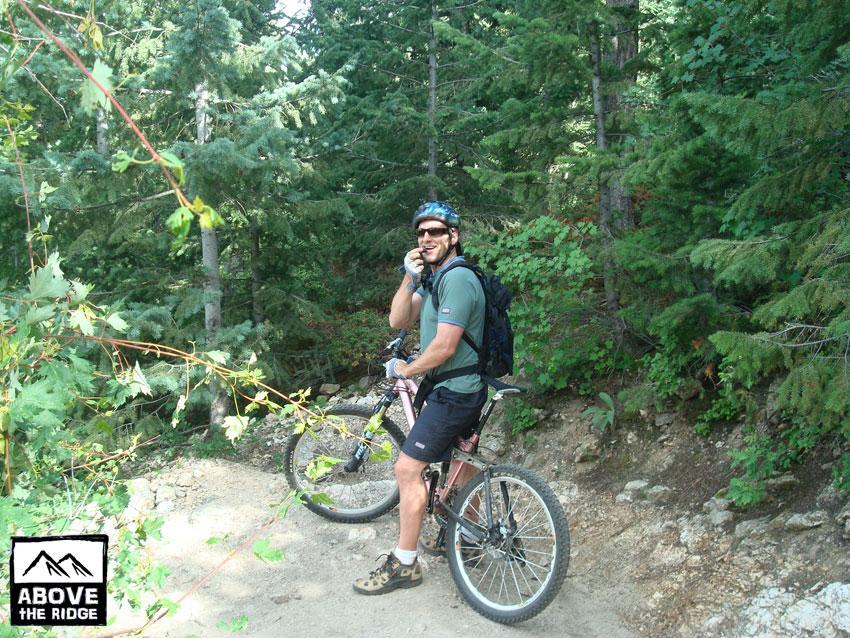A mountain biker wearing a helmet and sunglasses, standing beside his bike on a forest trail. He is smiling and holding a phone, surrounded by lush green trees and foliage. The image includes a logo for "Above the Ridge" in the bottom left corner. Elk Meadow mountain bike trail.