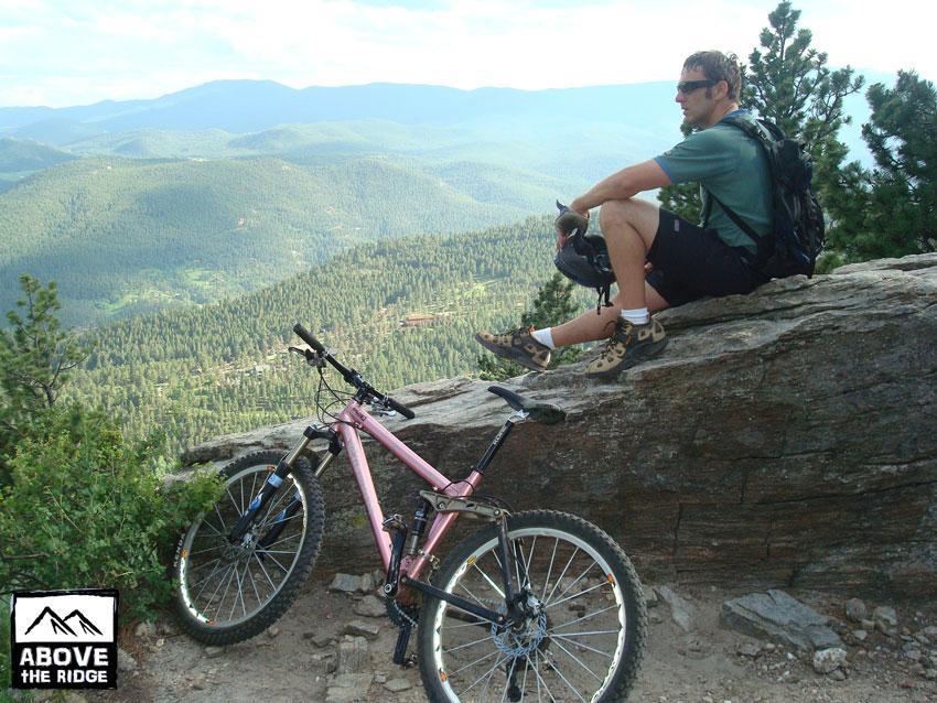 A mountain biker sitting on a rock overlooking a scenic vista of lush green mountains and valleys, with a pink mountain bike leaning against the rock nearby. The rider is wearing sunglasses, a helmet, and a backpack, enjoying a moment of rest in nature. Elk Meadow mountain bike trail.