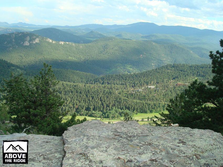 A panoramic view of a lush green landscape featuring rolling hills and dense forests under a blue sky with scattered clouds. The foreground includes rocky outcrops and evergreen trees, while the background showcases distant mountains. Elk Meadow mountain bike trail.