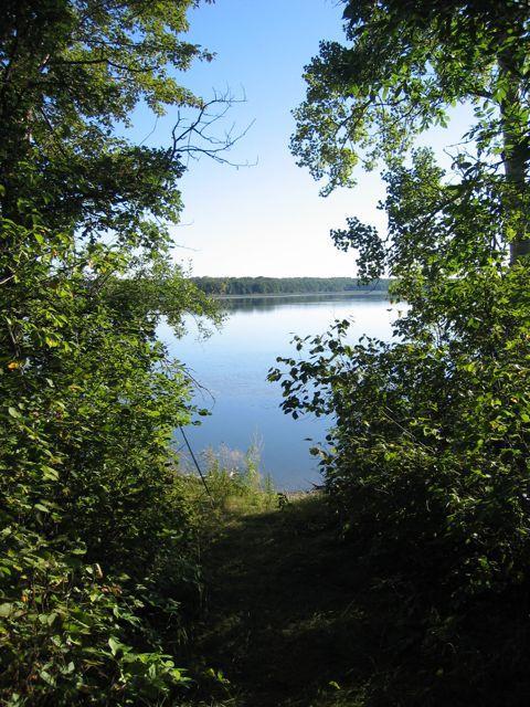A serene view of a calm lake framed by lush greenery. The scene captures a path leading through trees to the water's edge under a clear blue sky. Lake Metigoshe State Park mountain bike trail.