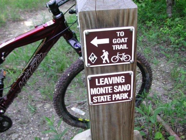 A wooden signpost with two signs. The upper sign points left towards "Goat Trail" with symbols for hiking and biking, while the lower sign states "Leaving Monte Sano State Park." A mountain bike is parked next to the sign, and the surrounding area features greenery and a dirt path. Monte Sano State Park &amp; Land Trust mountain bike trail.