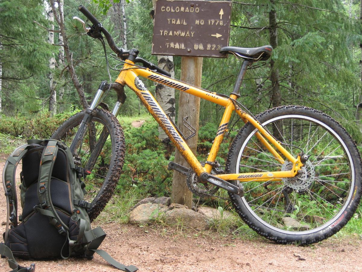 A yellow mountain bike with a muddy frame and tires leans against a wooden trail sign in a forested area. The sign indicates directions to Colorado and various trails, while a dark green backpack rests on the ground beside the bike. The surroundings include lush green trees and a dirt path, suggesting a nature trail environment. Colorado Trail: Green Mountain mountain bike trail.