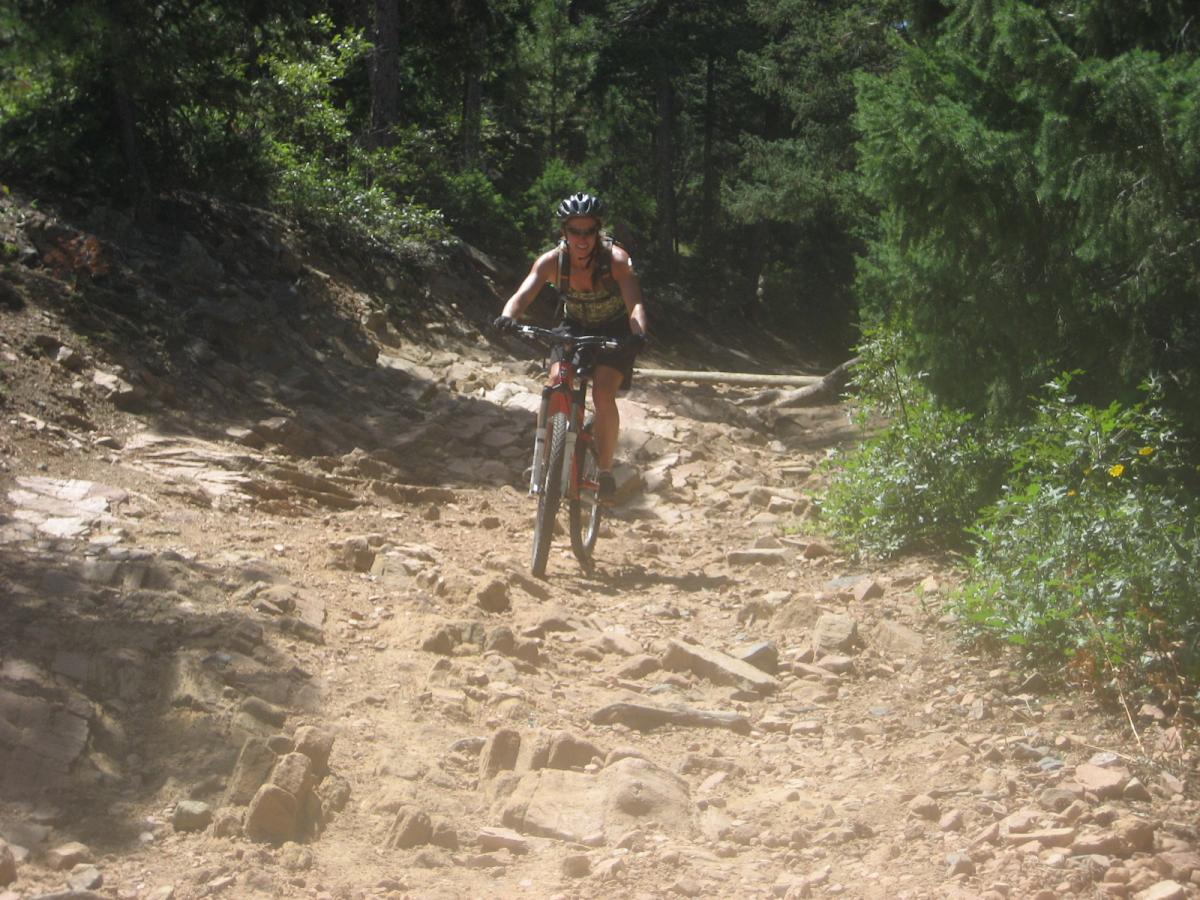 A mountain biker navigating a rocky trail surrounded by trees in a forested area. The rider is wearing a helmet and biking gear, showing focus as they tackle the uneven terrain under bright sunlight. Deer Creek Canyon mountain bike trail.