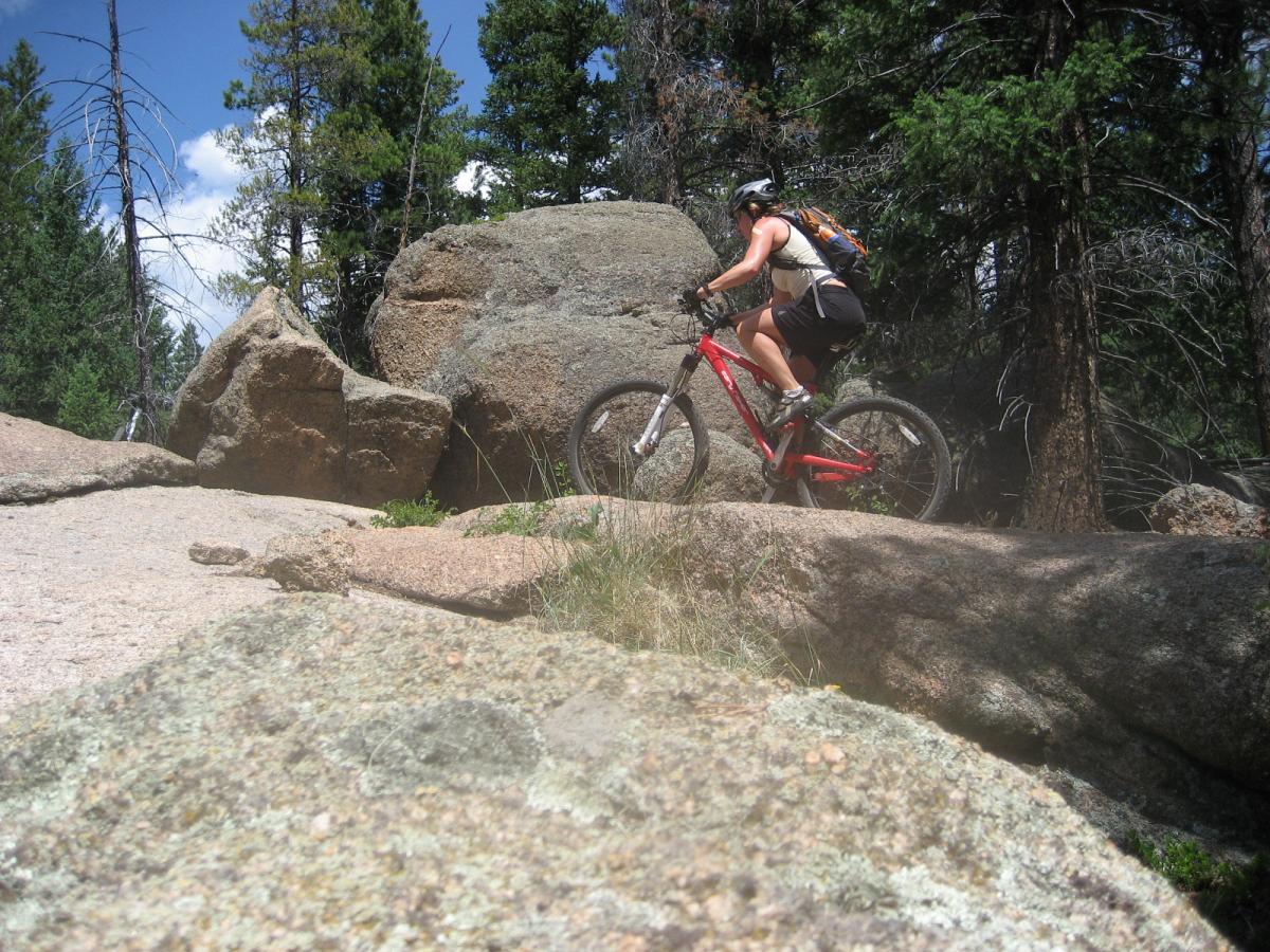 A mountain biker navigating rocky terrain in a forested area, surrounded by trees and blue sky. The rider is on a red bike, wearing a helmet and a backpack, and is positioned on a large boulder with smaller rocks and grass in the foreground. Colorado Trail: Green Mountain mountain bike trail.