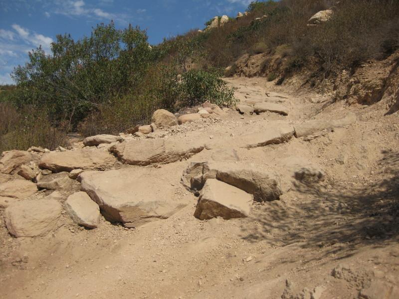 A rocky dirt trail winding uphill, surrounded by sparse vegetation and clear blue skies. The path is uneven with large stones and loose dirt, suggesting a moderate hike through a natural landscape. Sycamore Canyon mountain bike trail.
