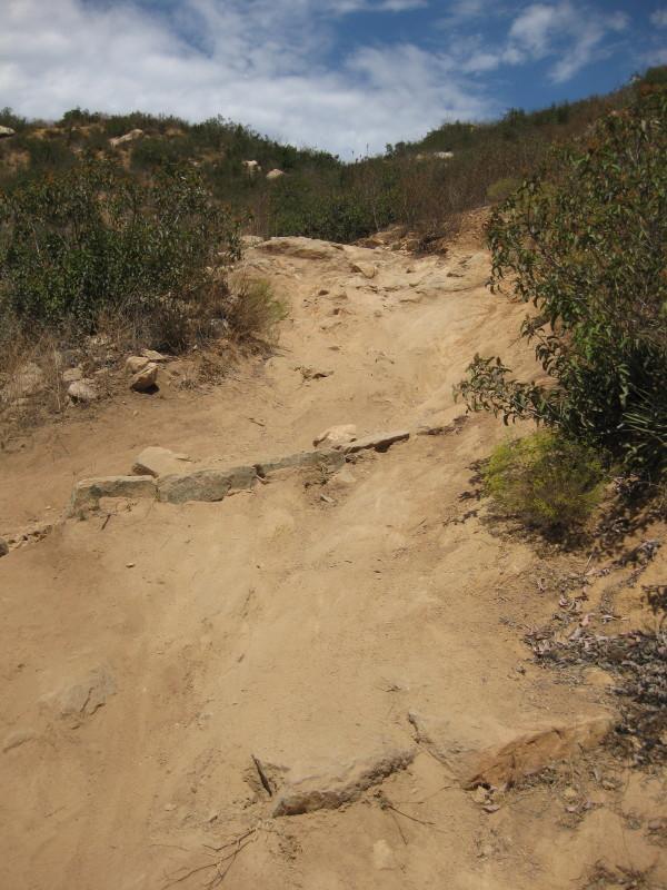 A rugged dirt trail leading uphill, flanked by sparse vegetation and rocky outcrops under a partly cloudy sky. The path appears uneven and steep, suggesting a challenging hike. Sycamore Canyon mountain bike trail.