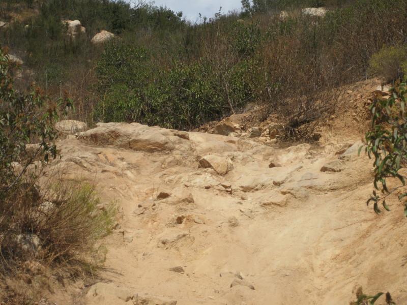 Rocky hillside path with sparse vegetation, featuring dry soil and scattered rocks, under a cloudy sky. Sycamore Canyon mountain bike trail.
