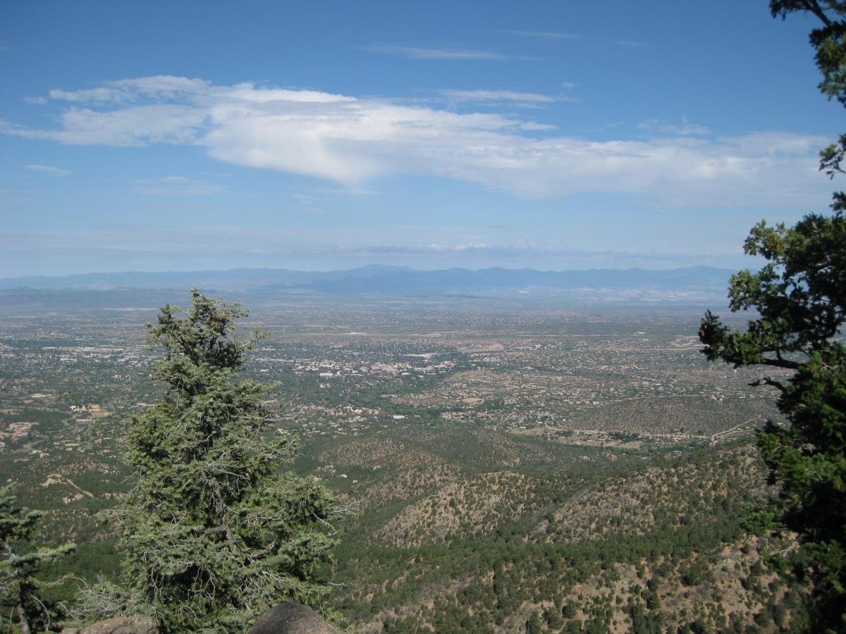 A panoramic view of a lush valley and distant mountains under a partly cloudy sky, with green trees in the foreground. Atalaya Mountain (174) mountain bike trail.