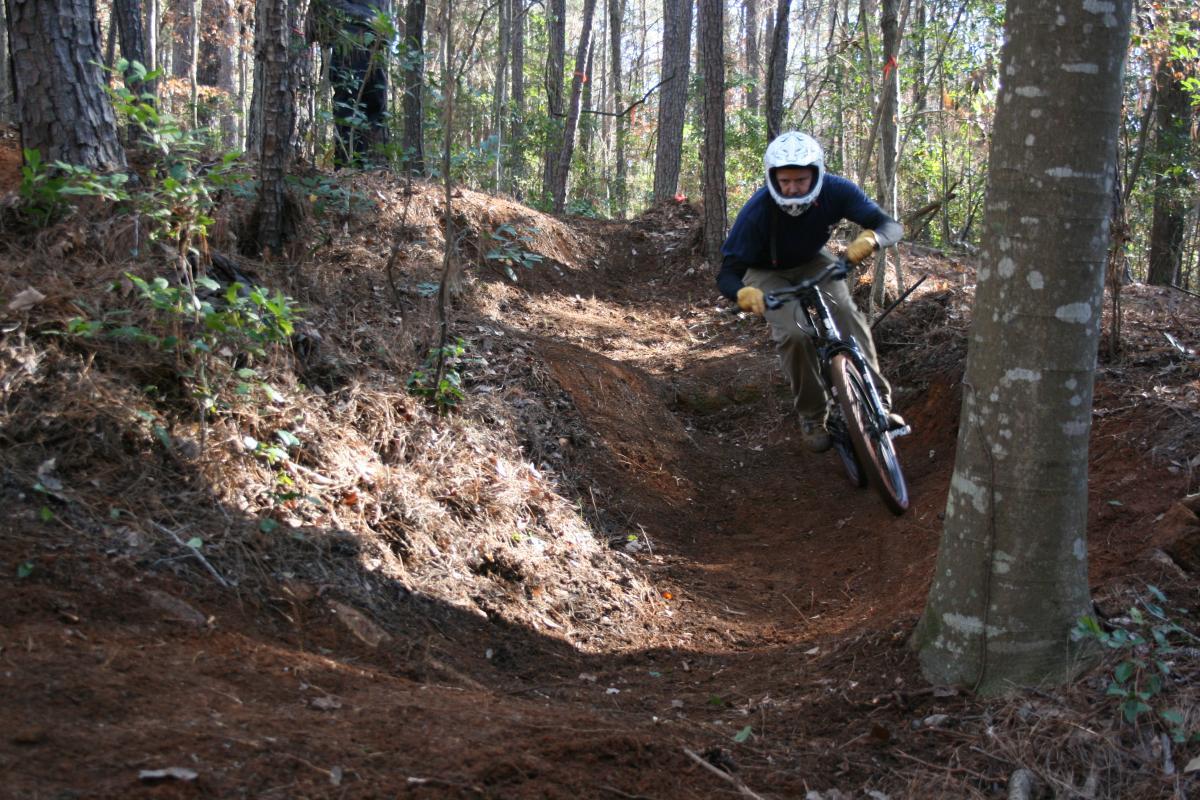 A mountain biker navigating a dirt trail in a wooded area, leaning into a turn while wearing protective gear and a helmet. The scene features tall trees and a forested landscape with sunlight filtering through the leaves. Raider Trails mountain bike trail.